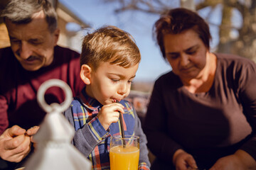 Small caucasian boy drinking juice at cafe or at home while sitting with his grandmother and grandfather in sunny day indoor - real people family leisure concept