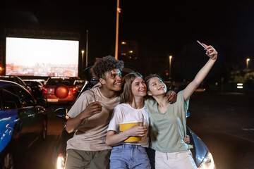 Joyful diverse young best friends posing while taking a selfie together, standing in front of a big screen, ready to watch a movie in an open air cinema