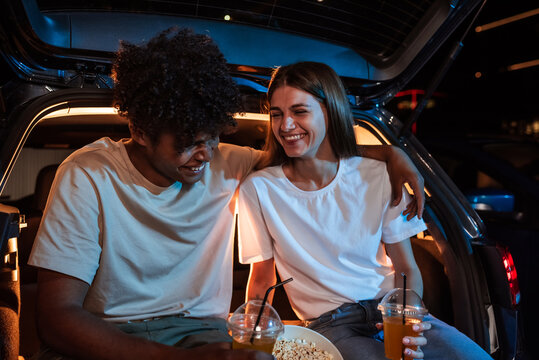 Portrait Of Diverse Young Couple Having Fun Together. Cheerful Girl And Her Boyfriend Laughing While Sitting Together In A Car, Parked In A Drive In Cinema