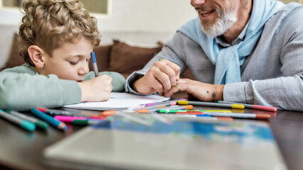Cute little boy drawing with colored felt tip pens on paper with cheerful grandfather, siting together at table in the living room