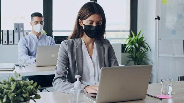 Video of two attractive business partners wearing a hygienic face mask while working with laptops in the coworking space. Social distancing concept.