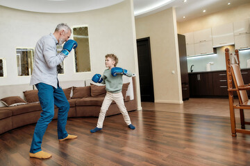 Mature man grandfather wearing boxing gloves practicing punches with little grandson. Physical activity for kids at home