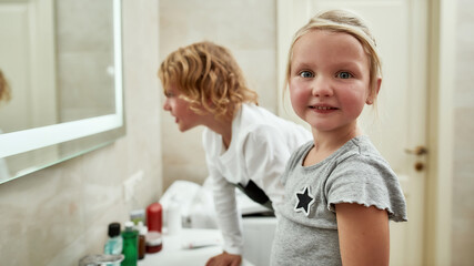 Portrait of cute little girl smiling at camera while washing her face, brushing teeth together with her sibling brother in the bathroom