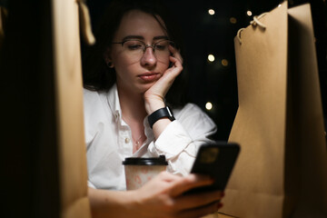 a young woman in glasses and a white shirt sits among paper bags with purchases, drinks coffee and looks at the phone