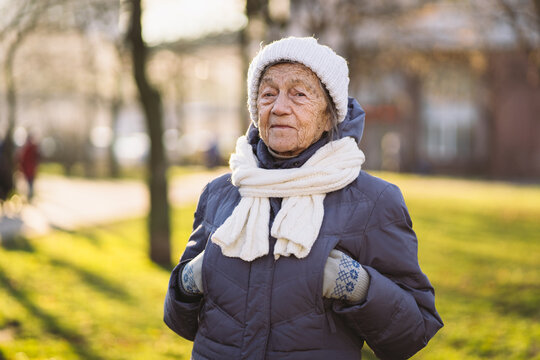 Portrait Caucasian Senior Woman With Gray Hair And Deep Wrinkles 90 Years Old Posing In Warm Clothes, White Knitted Scarf And Hat In Park, Sunny Frosty Weather. Active Old Age, Walking Elderly Female