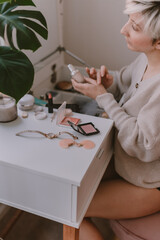 Young millennial woman is dressing up. Wearing earrings. Beauty routine. Make up artist. Mascara, lipstick, blushes. Pastel colors. Dressing vanity mirror. Pretty blonde lady. White bedroom light.