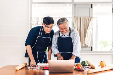 Portrait of happy love asian family senior mature father and young adult son having fun cooking together and looking for recipe on Internet with laptop computer to prepare the yummy eating lunch 