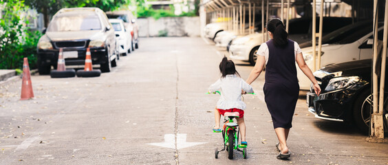 Children learns to ride a bike. Mother helped her daughter hold the bicycle. Mom helped guide the bike to the little child girl. Kid learns to ride a bicycle against counter car lens. Rear view people