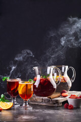 On the table, Two jugs with lemonade, mulled wine and glasses filled with drinks. Steam develops from drinks. Strawberries, apeltsins, raspberries and sprigs of fresh mint are on the table