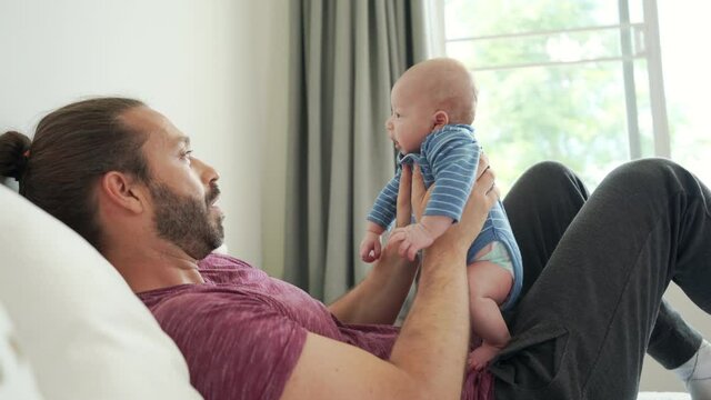 Happy family stay home. Smiling handsome Caucasian man carrying and playing with his adorable newborn baby son on the bed in bedroom. Father caring cute infant child boy and relaxing together at home.