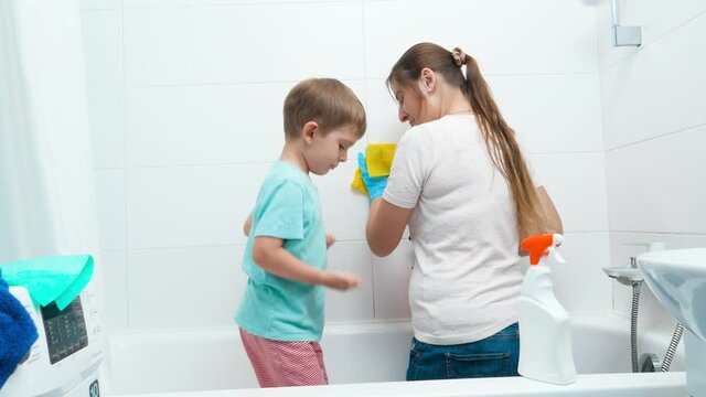 Funny toddler boy dancing and having fun while mother doing housework and washing walls in bathroom
