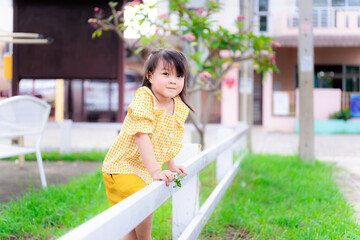 Fototapeta premium Little girl in a yellow dress stood by the white fence. Child pretended to be climbing the fence to the other side. Children smiled sweetly and looked at the camera. Kid are 5 years old.
