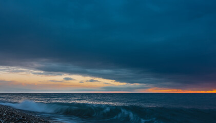 Evening at the sea in blue tones. Dense clouds cover almost the entire sky. The open area is illuminated by the setting sun in orange. The wave foams on the beach pebbles. Black Sea