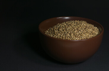 Brown uncooked buckwheat in a bowl with scattered grains nearby on a black background