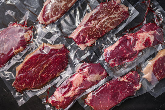 Set  of  vacuum packed organic raw beef alternative cuts: top blade, rump, picanha, chuck roll steaks, over black textured background, top view.