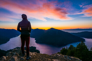 Adventurous athletic woman hiker standing on a mountain top watching the sunset over mountains and a beautiful lake.