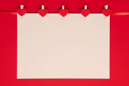 Blank sheet of paper on clothespins with hearts on red background. Backdrop for greeting or invitation to valentine's day, wedding. Romantic mock up.