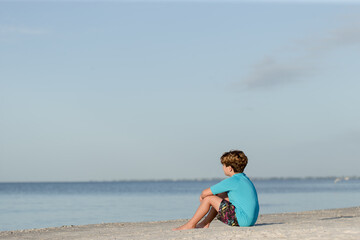 Young boy sitting on beach looking out over water like contemplating his future isolated depressed and alone