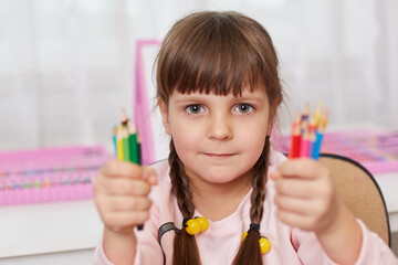 Adorable little girl with lots of multicolored pencils in hands posing indoor, looking directly at camera with serious look, asks for help to decide what pencil to choose.