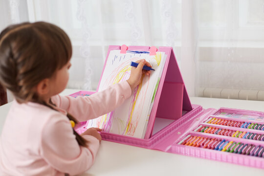 Little girl with dark hair drawing with colourful pencils on canvas, has great variety of multicolored pencils and wax crayons, kid sitting at table in front of window.
