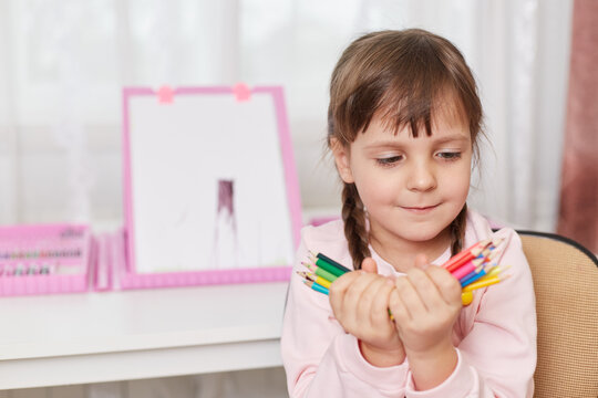 Portrait of child girl holding colorful pencils in both hands and looking at it, small lady with pigtails decides what colour to choose for painting her picture.