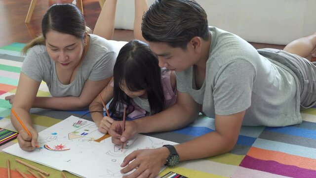 Happy Asian family at home. Parents with little daughter lying on the floor using color pencil drawing on paper book. Smiling father and mother with cute girl kid having fun homeschooling together.