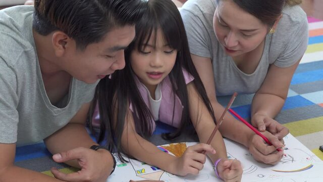 Happy Asian family at home. Parents with little daughter lying on the floor using color pencil drawing on paper book. Smiling father and mother with cute girl kid having fun homeschooling together.