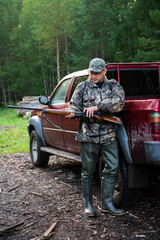 Man hunter standing with shotgun rifle near his car outdoor preparing for bird hunt in autumn forest