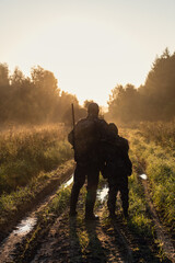 Hunters with hunting equipment going away through rural field towards forest at sunset during hunting season in countryside.