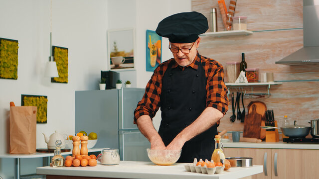 Baker kending dough in kitchen table wearing apron and bonete. Retired elderly chef with uniform sprinkling, sieving sifting raw ingredients by hand baking homemade pizza, bread.