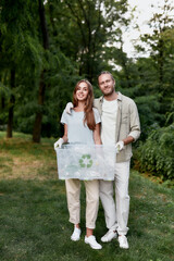 Fototapeta premium Vertical shot of young happy couple, eco volunteers holding recycle bin and looking at camera while collecting plastic waste together in the green forest