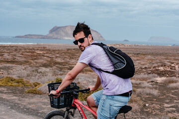 A young caucasian man with sunglasses riding a bike in a dirty road in La Graciosa, Canary Islands