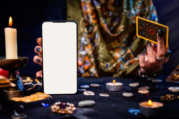 The fortune teller is holding a Tarot card and a smartphone with a white screen. Mock up. Close-up....