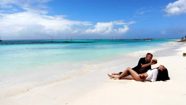 Exotic relax at Maldives beach, young couple in love in bikini lying on whie sand and tasting tropical beverages. Happy people enjoying summer vacations, discovering exotic places. Love emotions