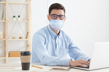 Young employee sitting in office, working with laptop, wearing mask, practicing social distancing because covid safety protocol