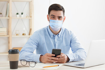 Young business man sitting in office, wearing mask, practicing social distancing because covid safety protocol