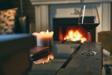 Glass of red wine with book and candle on table at home, fireplace in the background. Warm, dark colors.
