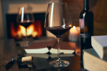 Two glasses of red wine with book and candle on table at home, fireplace in the background. Warm,...