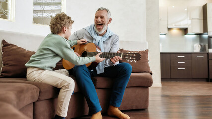 Weekend with grandpa. Cheerful mature man, grandfather playing guitar to his cute little grandson