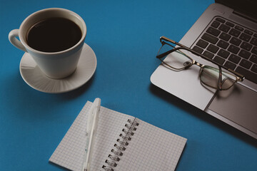 Desktop workspace with laptop, eyeglass, cup of coffee and notes bloc. Blue background. 