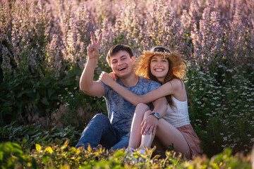 Fototapeta premium Happy couple in love sits hugging by blooming sage field. A young man with beautiful girl in straw hat are having fun, fooling around, waving their hands, showing their tongue. travel of crazy lovers