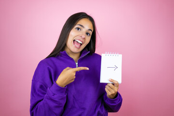 Fototapeta na wymiar Young beautiful woman wearing sweatshirt over isolated pink background smiling holding notebook with arrow message
