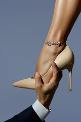 Male hand is holding female leg in beige high-heeled shoe. The female ankle is adorned with silver anklet with insert made as word "Nasty" decorated with a plenty of sparkling crystals. 