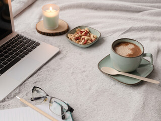 Coffee, biodegradable eco spoon, working from home concept, reading glasses, notebook, goji berries in a green bowl with cashews nuts, laptop computer, grey soft cozy hygge plaid blanket