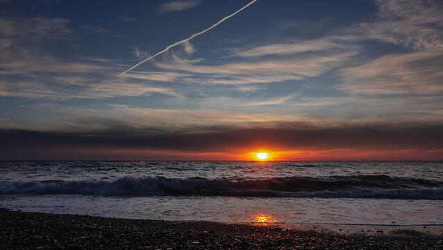 Bright sunset over the sea. In the blue sky there are clouds and an airplane track. The sun is low on the horizon. The wave rolls onto the pebble beach. Sunny path.