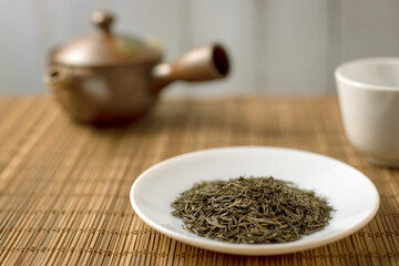 Closeup of dried organic green tea leaves on white plate (saucer) put on Japanese style bamboo wooden placemat with teapot and cup in background 