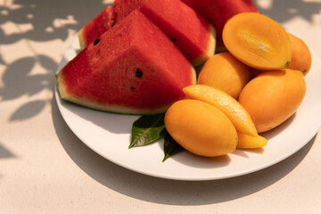 Closeup Plate of fresh fruits watermelon slice and plum mango or gandaria on a sandy background taken outdoors under natural lighting with shadow from a branch of leaves. Healthy eating concept