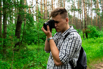 Boy, a male tourist take camera, photos in a gray-and-white checked shirt cap with a gray backpack, walking along a path in the forest, turning his head, looking into the distance. Tourist with a map