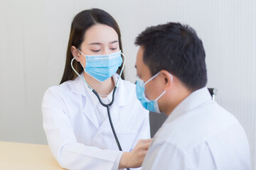 Asian woman doctor uses stethoscope to check up health of the man patient while both wear medical face mask to prevent respiratory system infectious from pathogens or Covid-19.