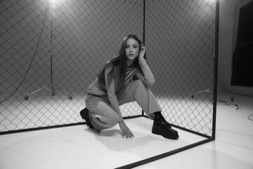 Obraz premium Fashion model in green sporty clothes and black boots sitting on white florr against grid fence and pink background. Girl with long straight brunette hair